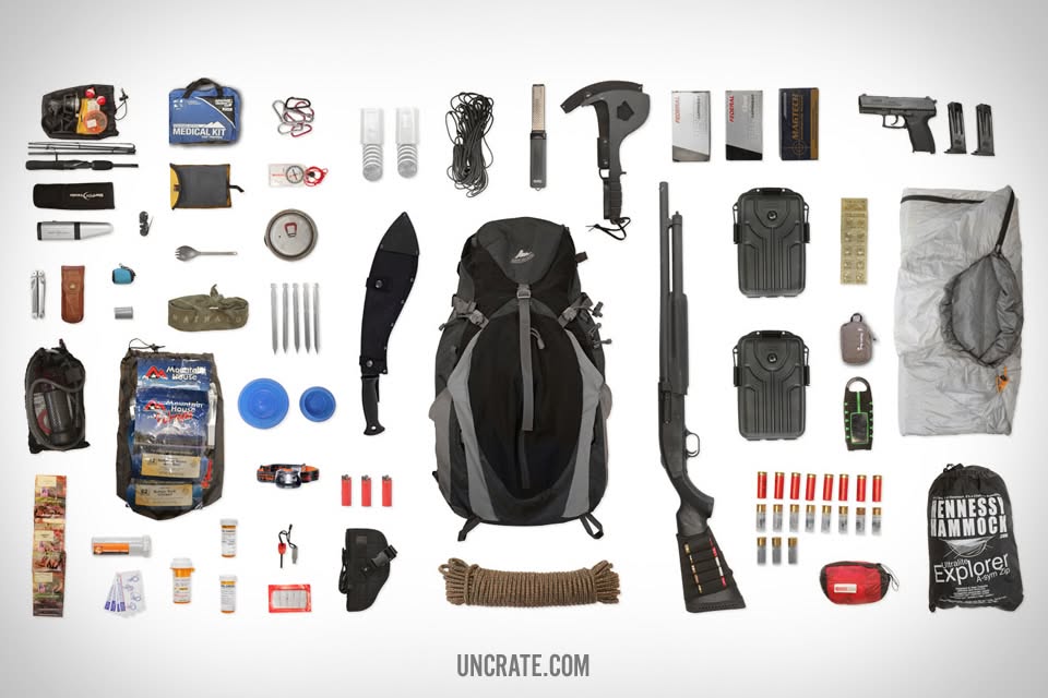 Survival gear laid out around a black backpack, including a medical kit, rope, a machete, a hatchet, food pouches, and a hammock